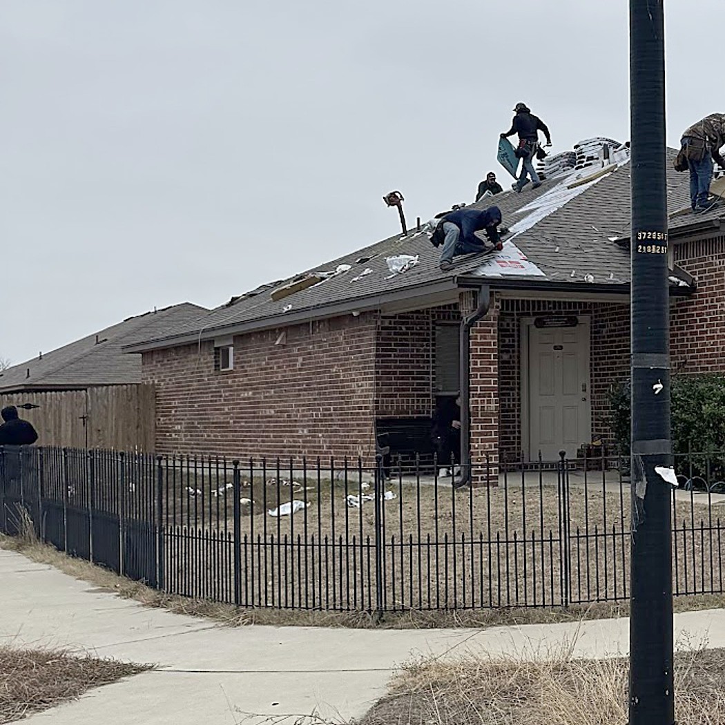 A crew working across the roof of a long single-story brick building behind an iron fence
