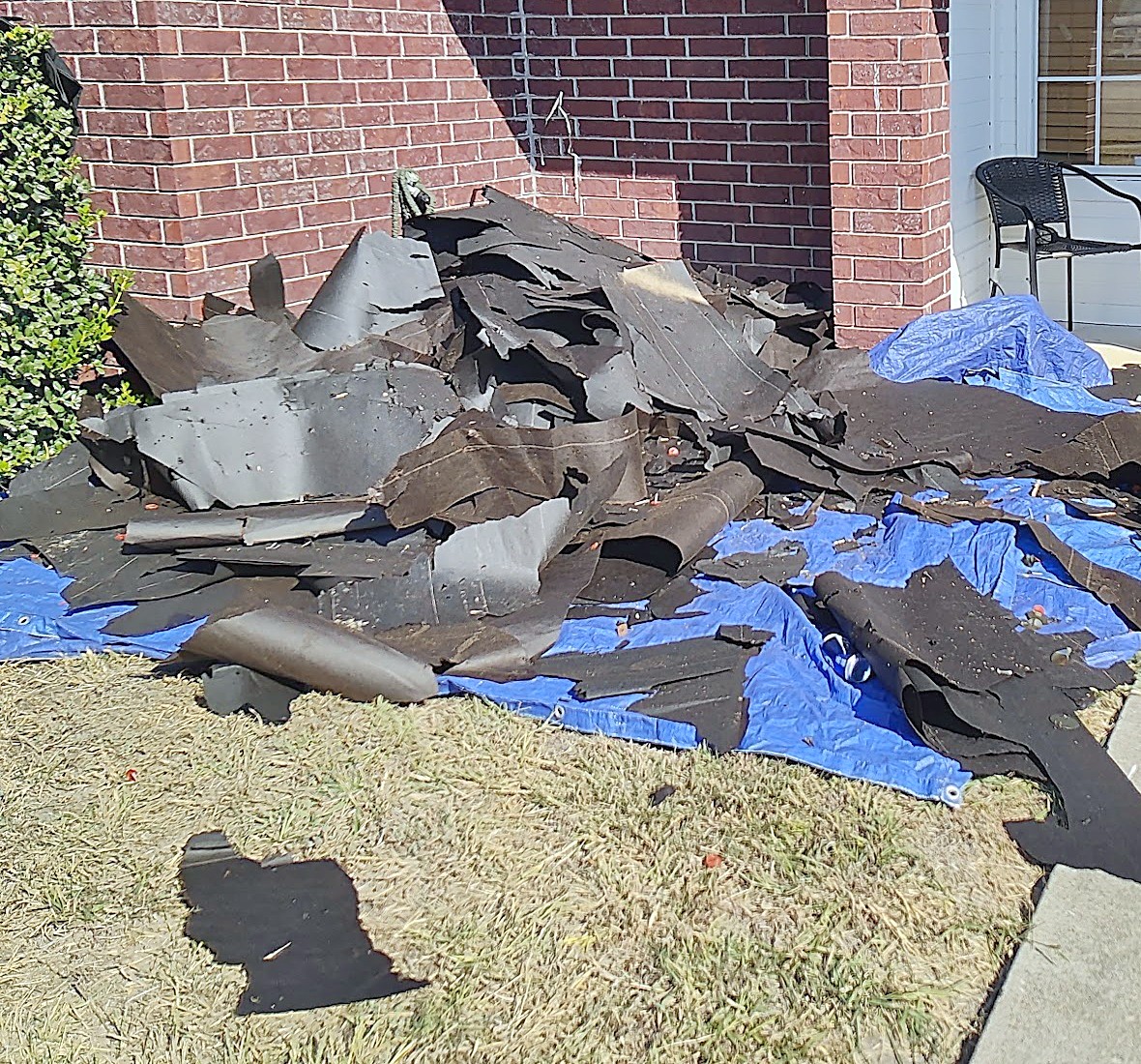 A pile of torn-off roofing spread across blue tarps laid on the ground beside a brick house