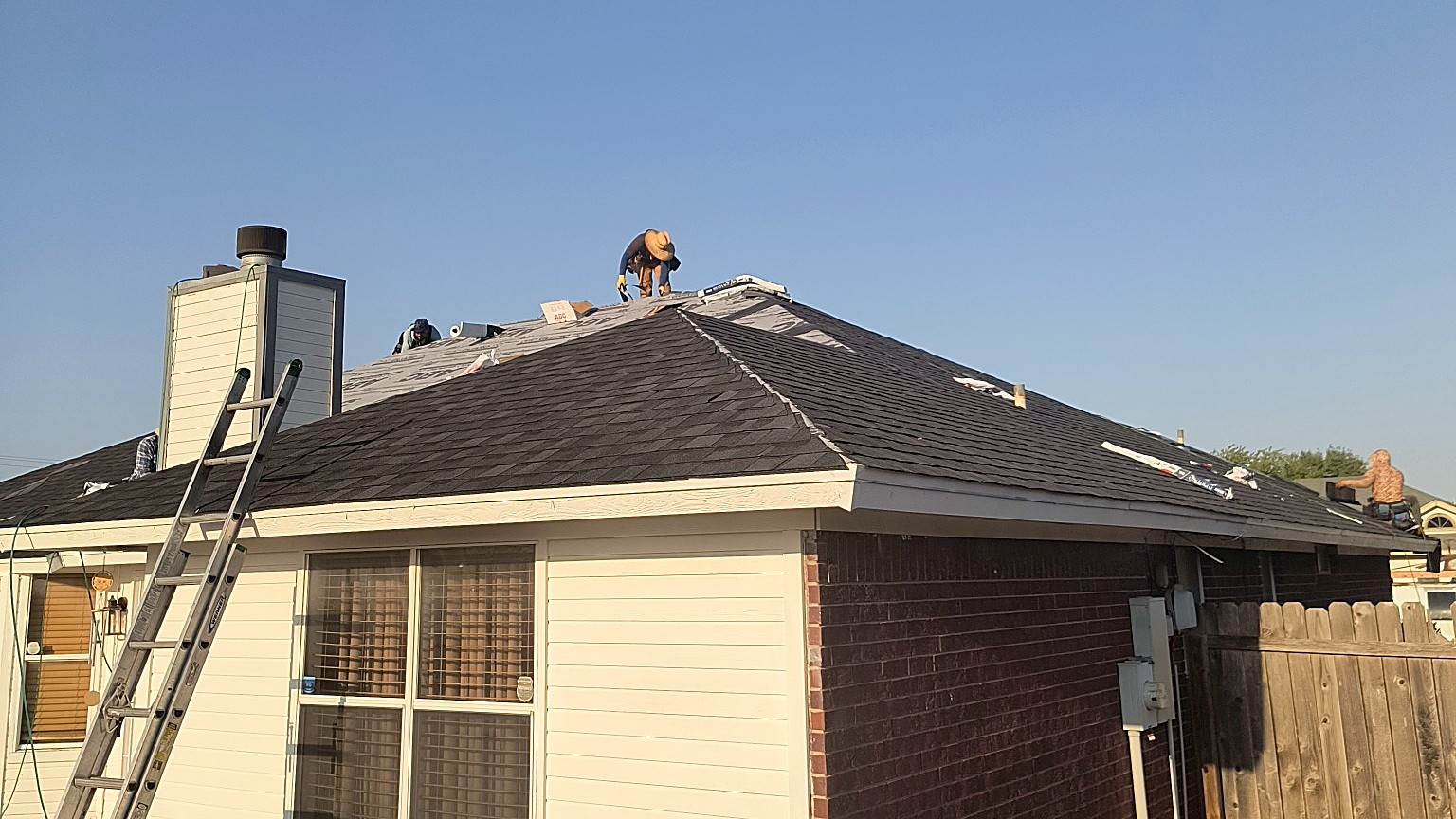 Three roofers laying shingles across the hip roof of a single-story house